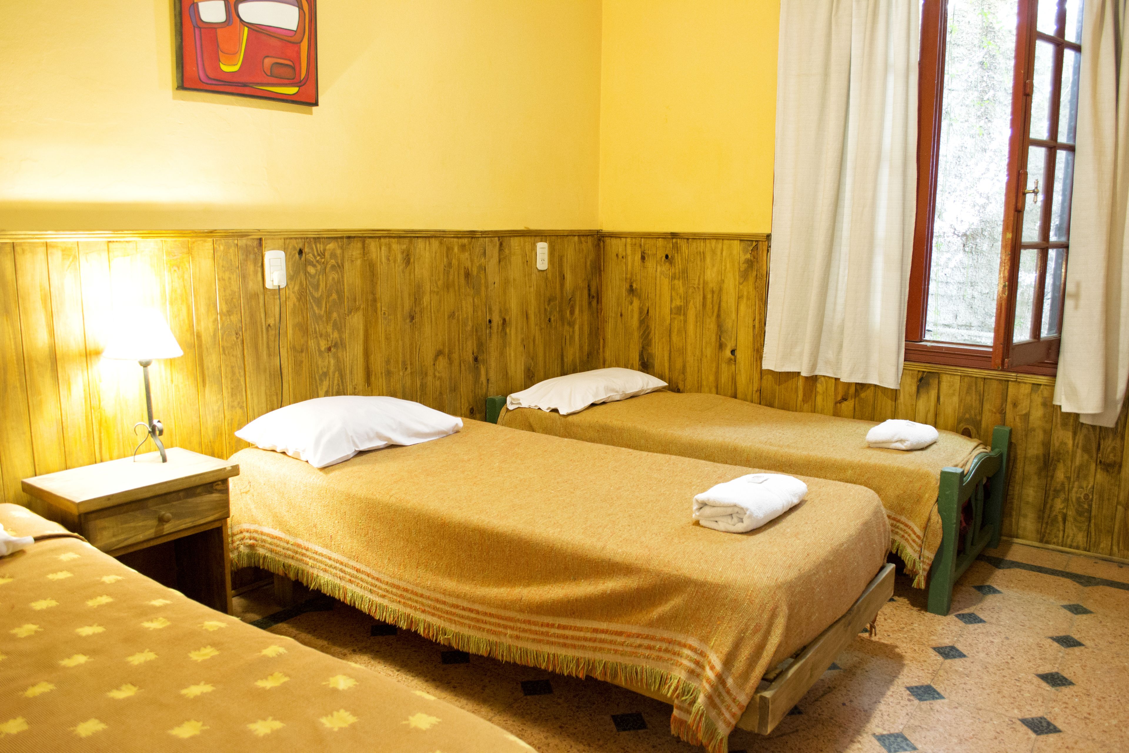 A room with two beds, wooden headboards, a nightstand with a lamp, and a window with curtains.