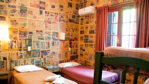 Una habitación con dos camas individuales, una litera de madera, una pared cubierta de periódicos, una ventana con cortinas y un equipo de aire acondicionado.