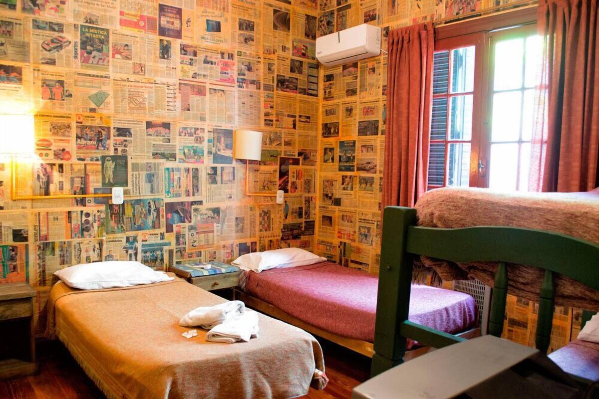A room with two single beds, a wooden bunk bed, a wall covered with newspapers, a window with curtains, and an air conditioning unit.