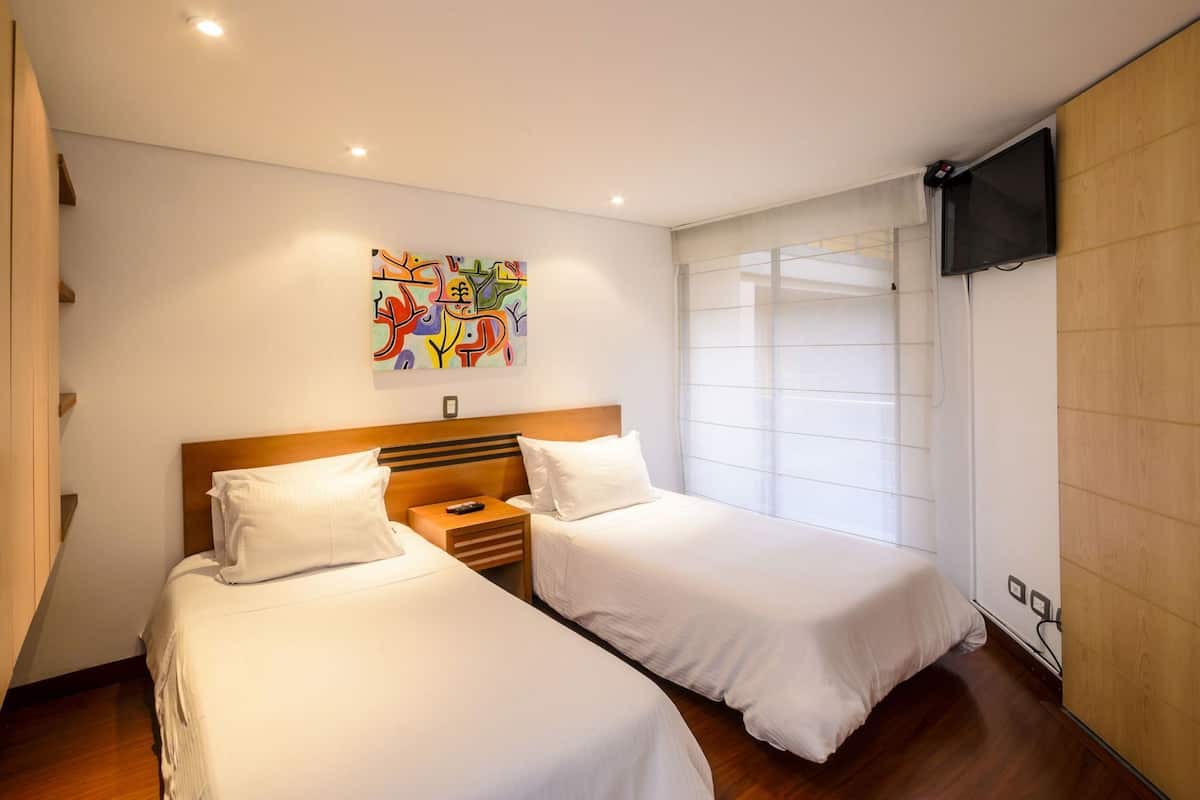 Une chambre d’hôtel avec deux lits, une tête de lit en bois, un tableau abstrait aux couleurs vives, un téléviseur fixé au mur et une fenêtre avec des stores.