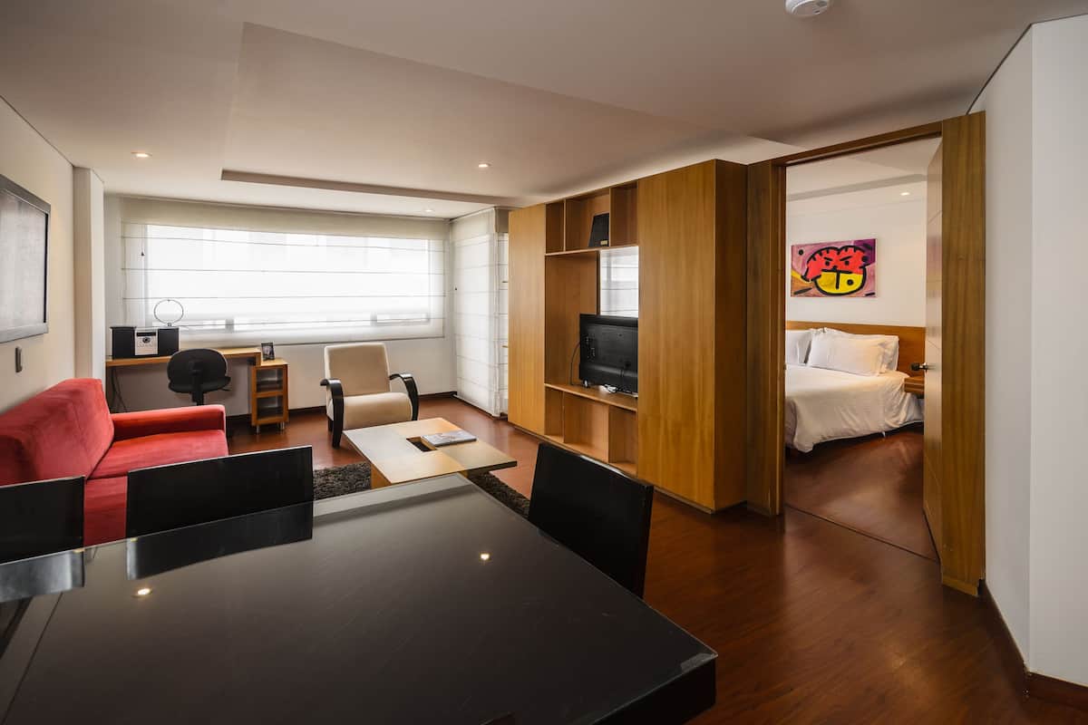 Une chambre d’hôtel moderne avec un coin repas, un canapé rouge, une télévision et un lit doté d’une tête de lit blanche.