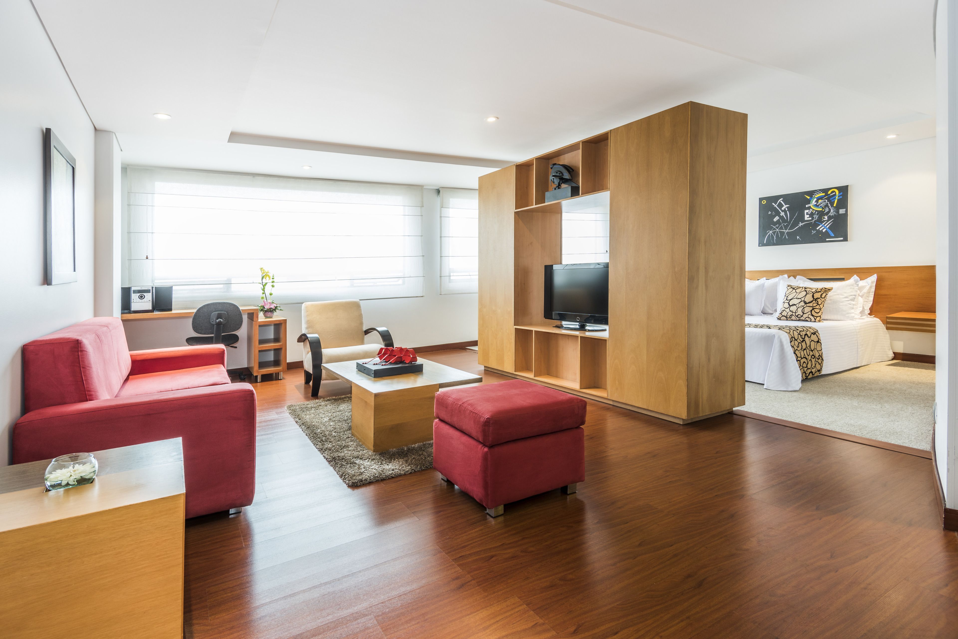 Ein modernes Hotelzimmer mit Holzboden, einem roten Sofa, einem kleinen Hocker, einem Couchtisch, einem Fernseher, einem Bett mit weißer Bettwäsche und einem großen Fenster mit Jalousien.