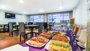A buffet setup with various fruits and beverages, arranged on a purple tablecloth in a room with wooden flooring and large windows. - Travelers Suites Obelisco Apartamentos (Bogotá)