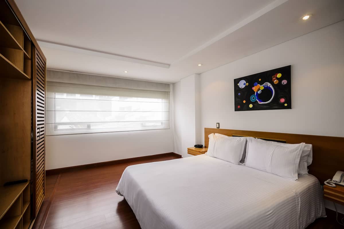 Une chambre d’hôtel avec un grand lit, une armoire en bois, une table de chevet avec un téléphone et une fenêtre avec des stores.