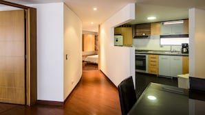 A modern kitchen with wooden cabinets, a refrigerator, and a coffee maker. There is a dining table with chairs and a hallway leading to a bedroom. - Travelers Suites Obelisco Apartamentos (Bogotá)