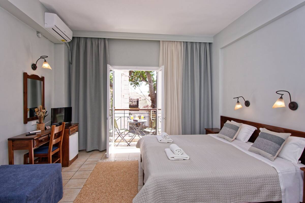 Een hotelkamer met een bed, bureau, stoel, televisie en een balkon met tafel en stoelen.