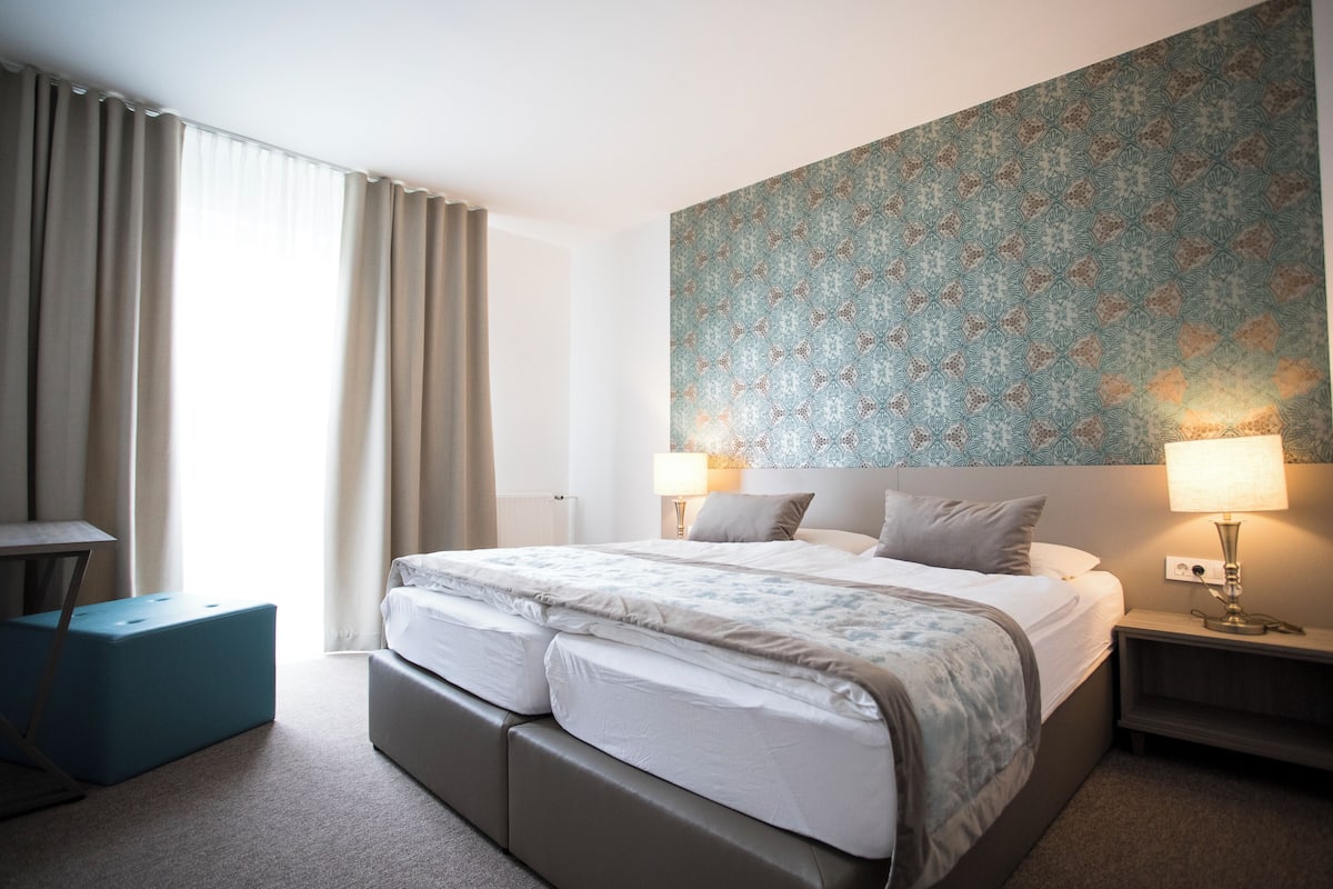 Habitación de hotel con una cama grande, dos mesitas de noche con lámparas, una mesita pequeña y un ventanal con cortinas.