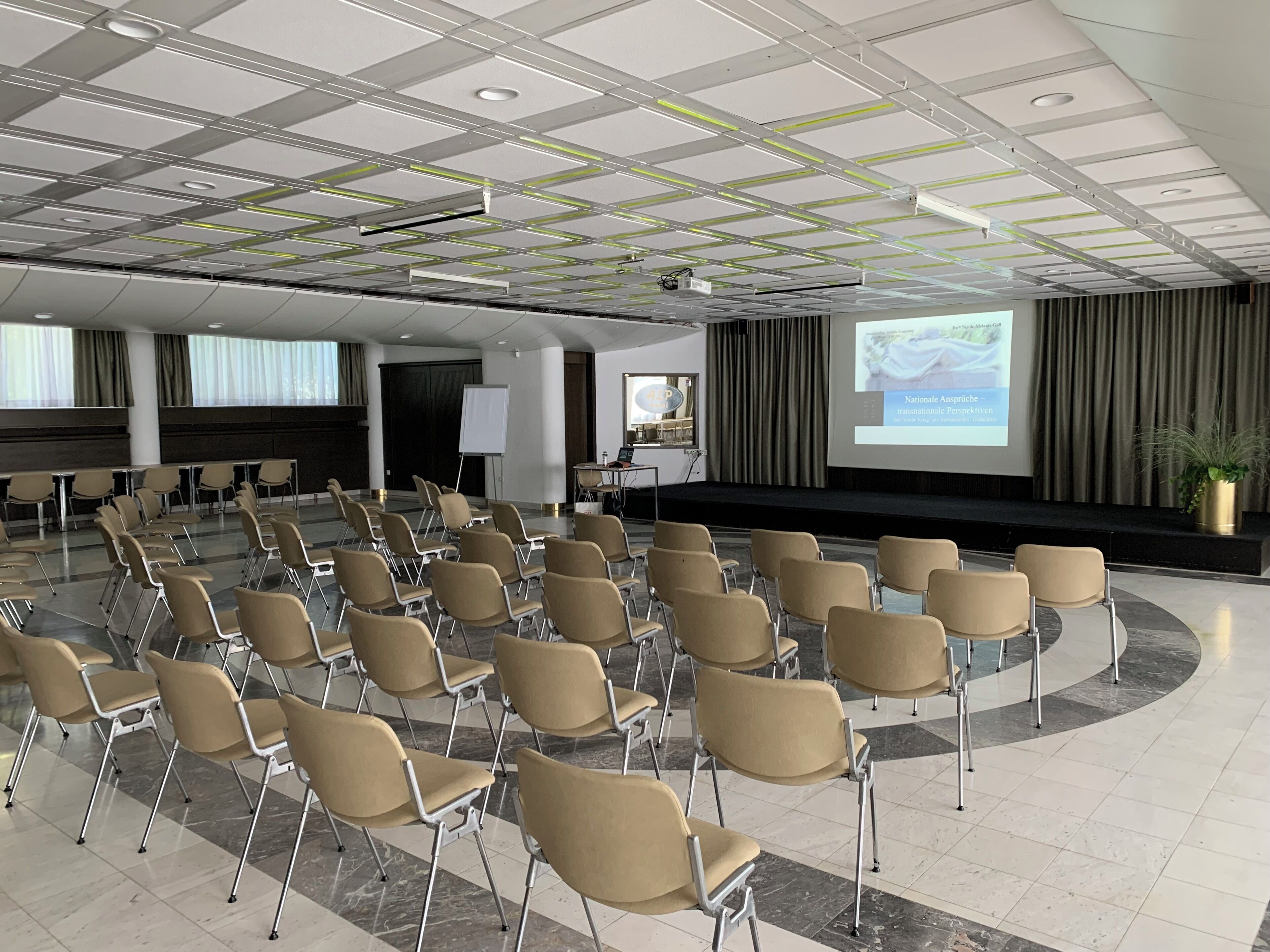 A conference room with rows of chairs, a stage with a projector, and a large screen displaying a presentation.