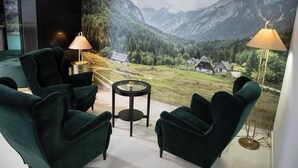 Habitación con vista panorámica a la montaña a través de un ventanal grande, con dos sillones verdes, una mesita redonda y dos lámparas de pie.