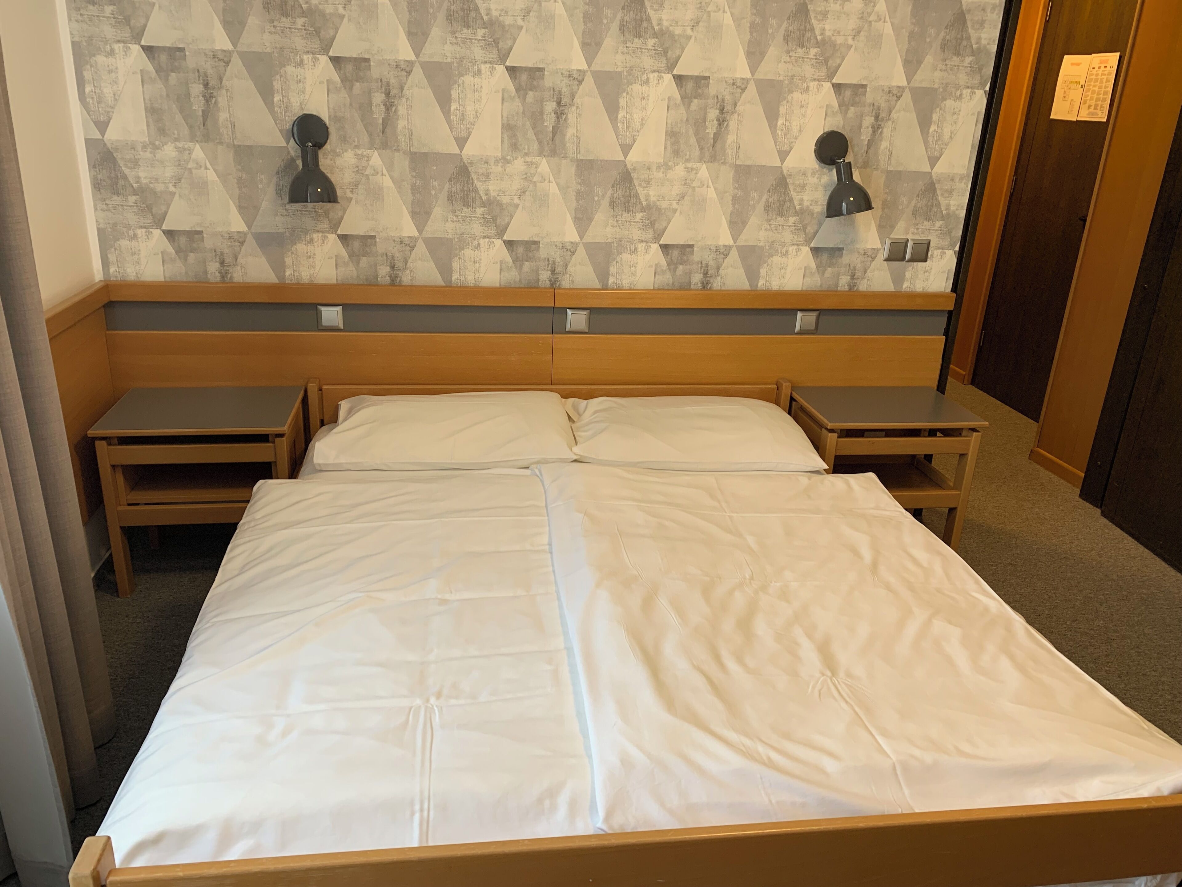 Cama doble con cabecera de madera, dos mesitas de noche y un pared con estampado geométrico.