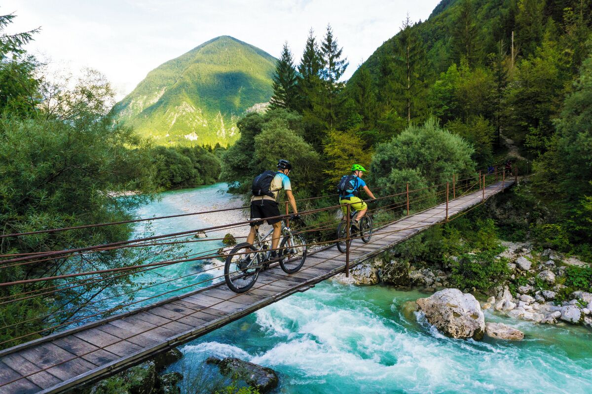 Two cyclists on a wooden bridge over a river with a mountainous backdrop.