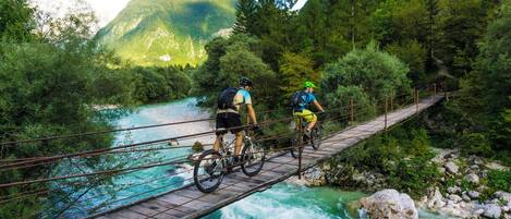 Two cyclists on a wooden bridge over a river with a mountainous backdrop.