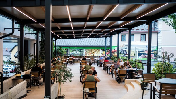 Un café al aire libre con mesas y sillas de madera, rodeado de plantas en macetas.