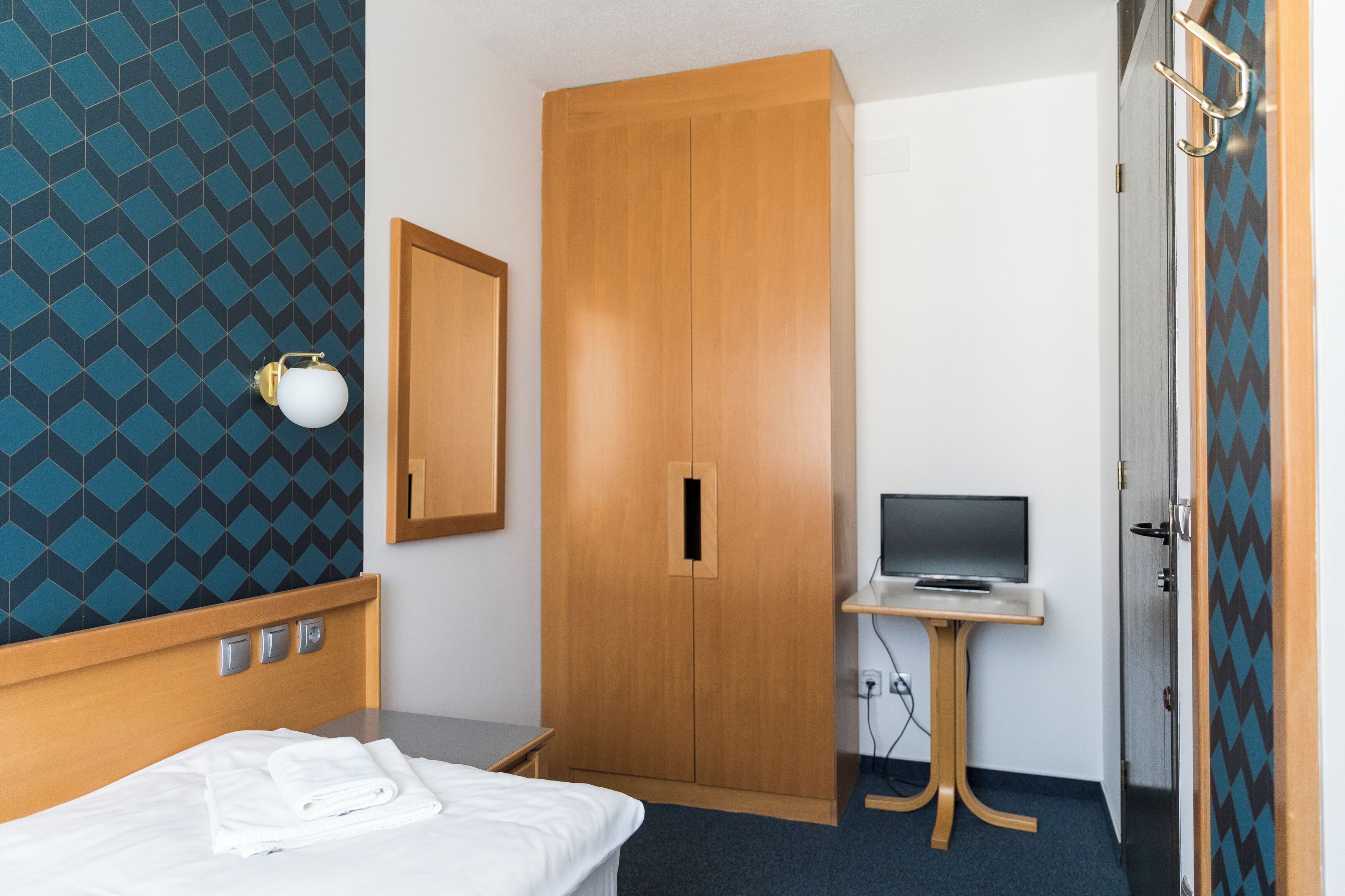 A hotel room with a bed, a wardrobe, a desk with a laptop, and a wall-mounted TV.