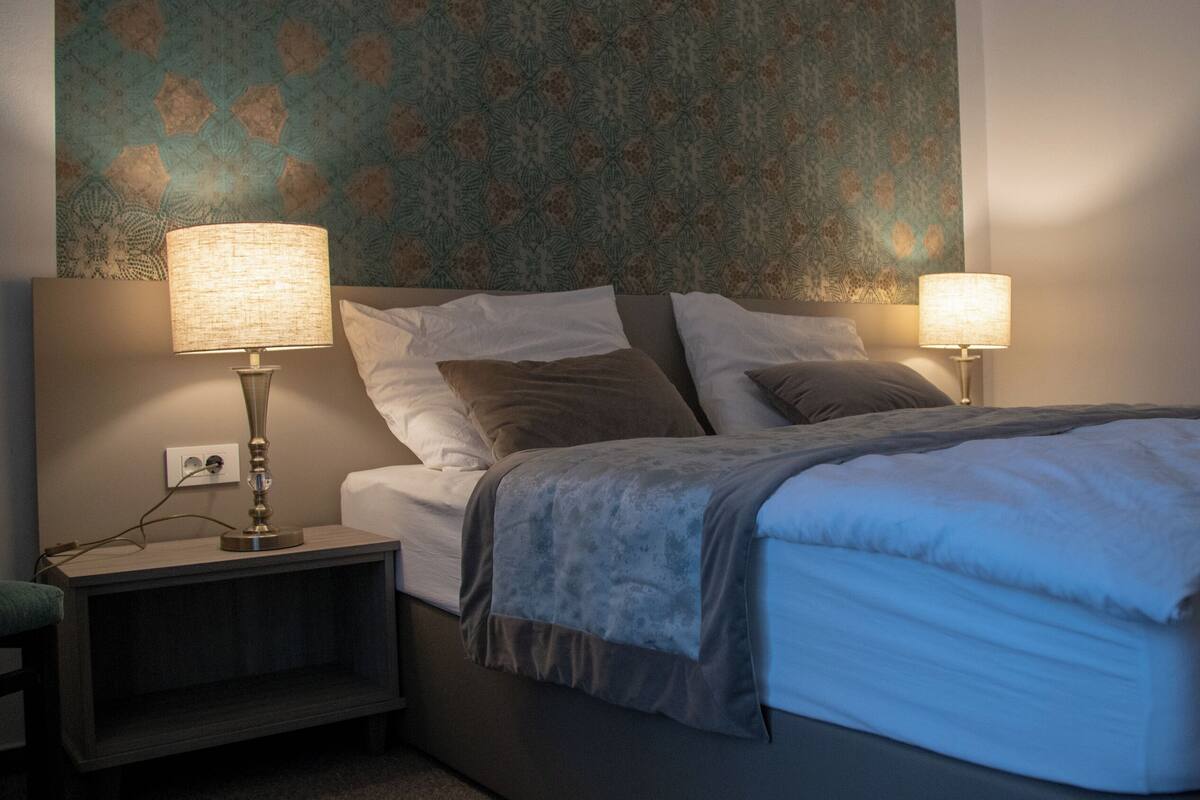 Una cama bien hecha con una manta gris, dos lámparas de noche, una mesita de noche con lámpara y una pared con diseño.