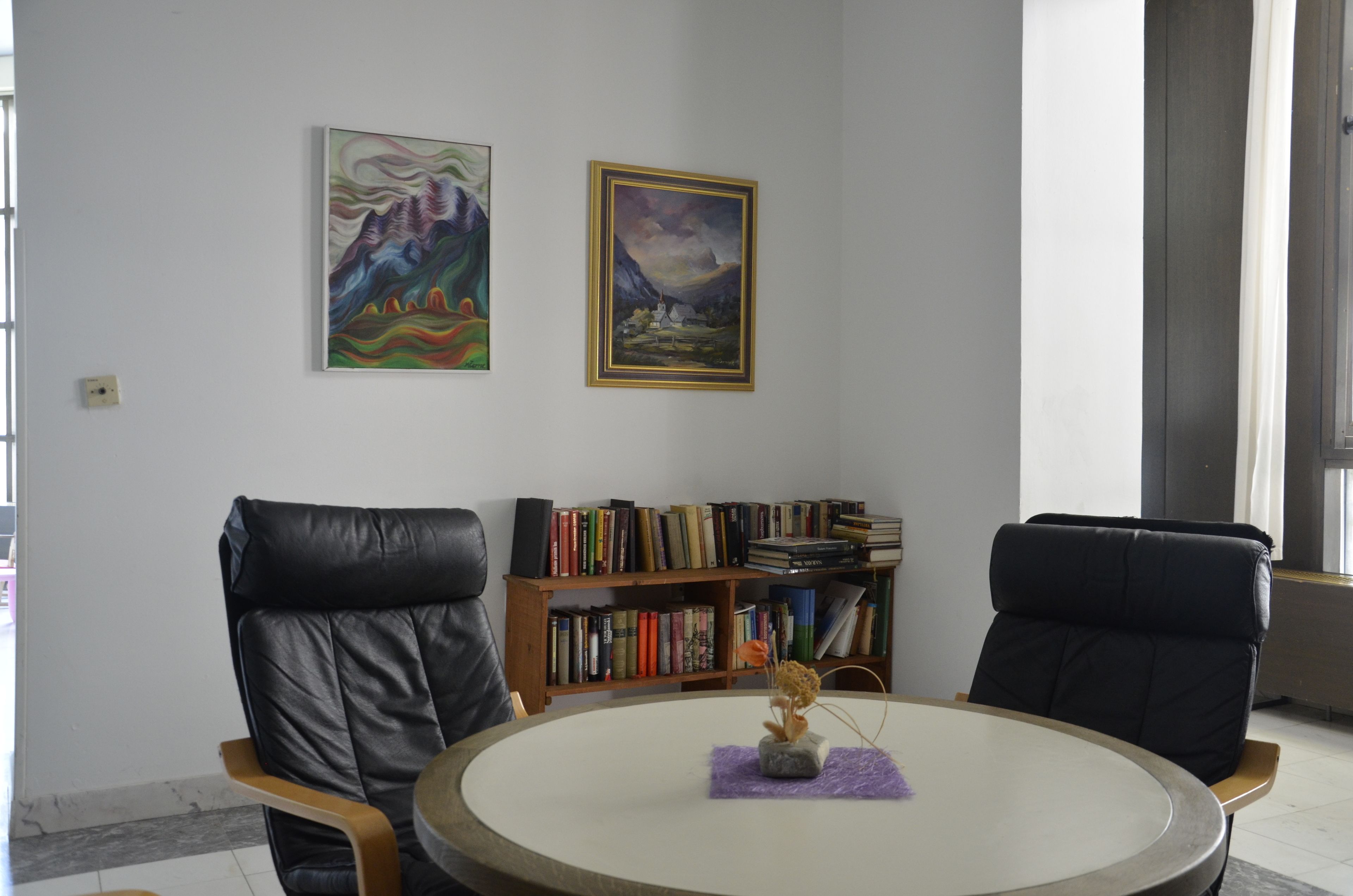 Quarto com duas poltronas de couro preto, uma mesa redonda com um vaso, uma estante e duas pinturas emolduradas na parede.