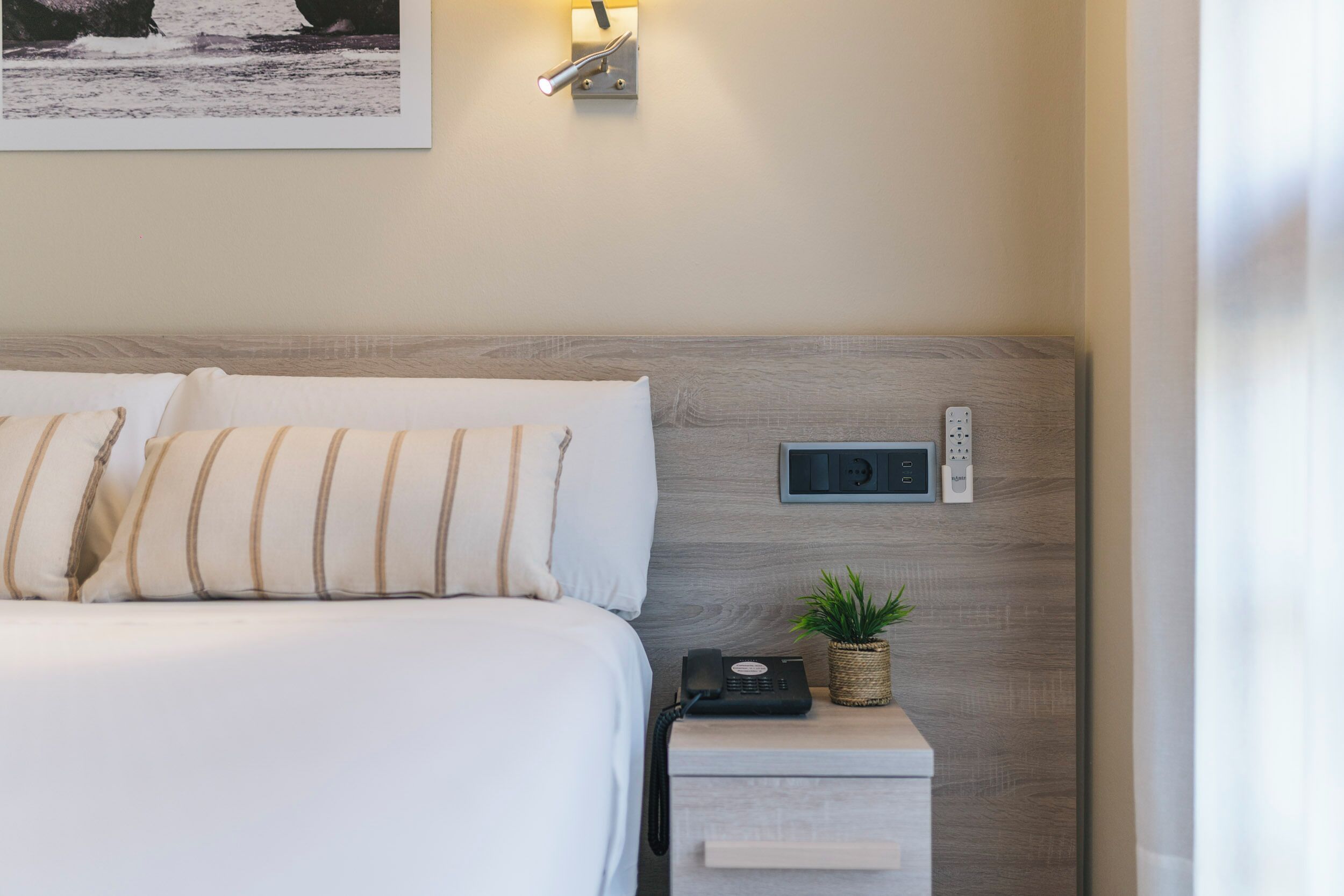 Un lit bien fait, agrémenté d’oreillers à rayures, une table de chevet avec un téléphone et une petite plante, et un luminaire fixé au mur.