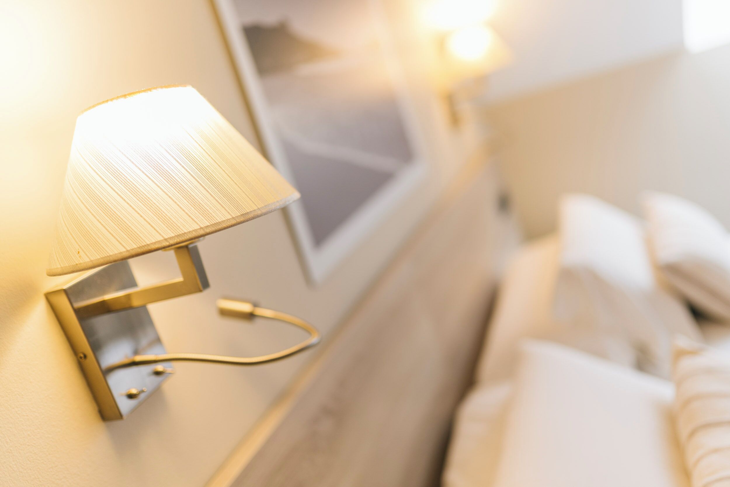 Een wandlamp met een metalen voet en een witte lampenkap, geplaatst in de buurt van een bed met witte lakens.