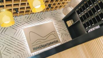 Interior de un hotel con piso estampado, un letrero que dice 'Hotel/Apartamentos' y un mueble de almacenaje negro con compartimentos numerados.