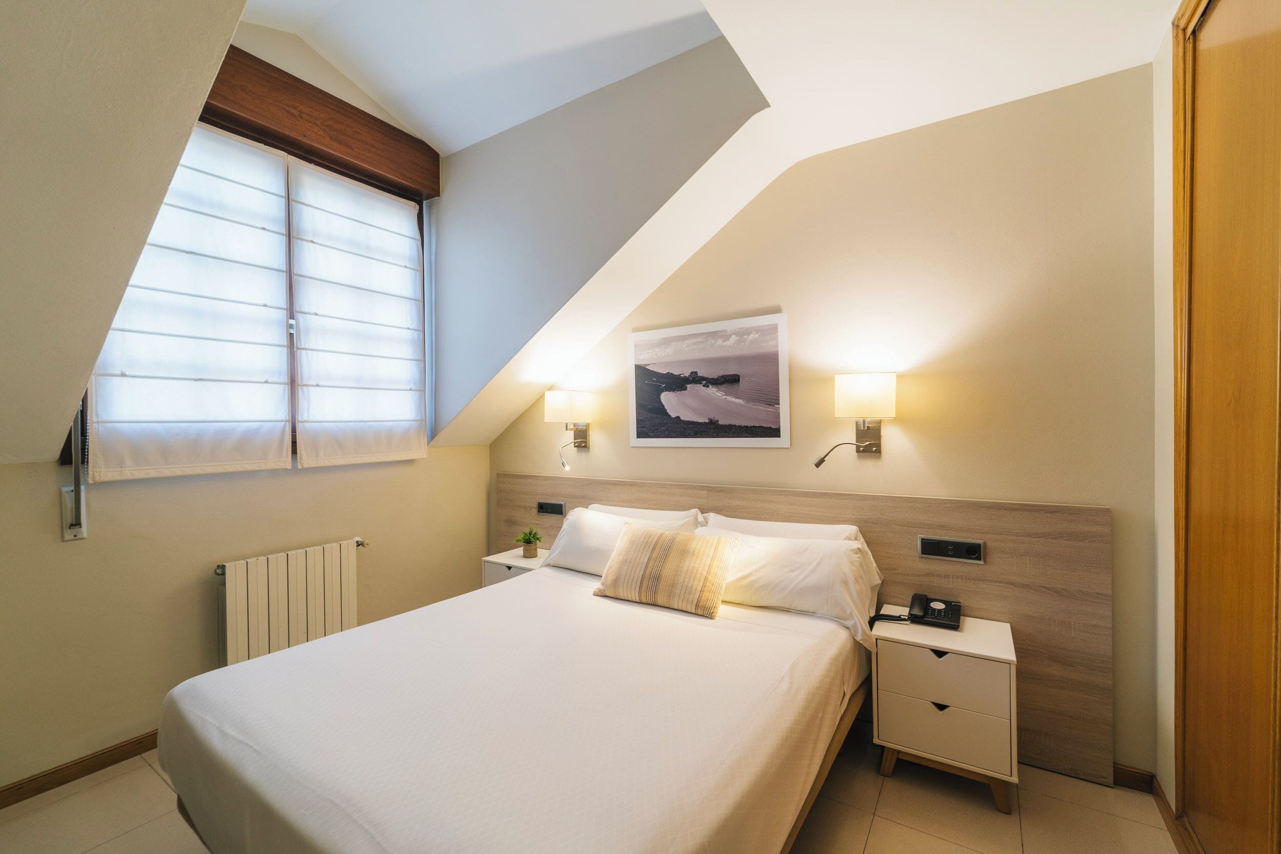 Un dormitorio con techo inclinado, una cama con ropa de cama blanca, dos mesitas de noche con lámparas, un radiador y una ventana con persianas.