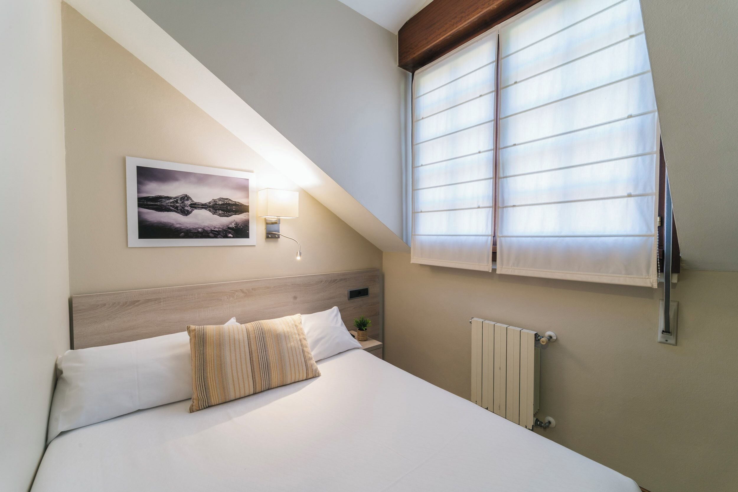 A bedroom with a bed, a nightstand, a wall-mounted lamp, a radiator, and a window with blinds.