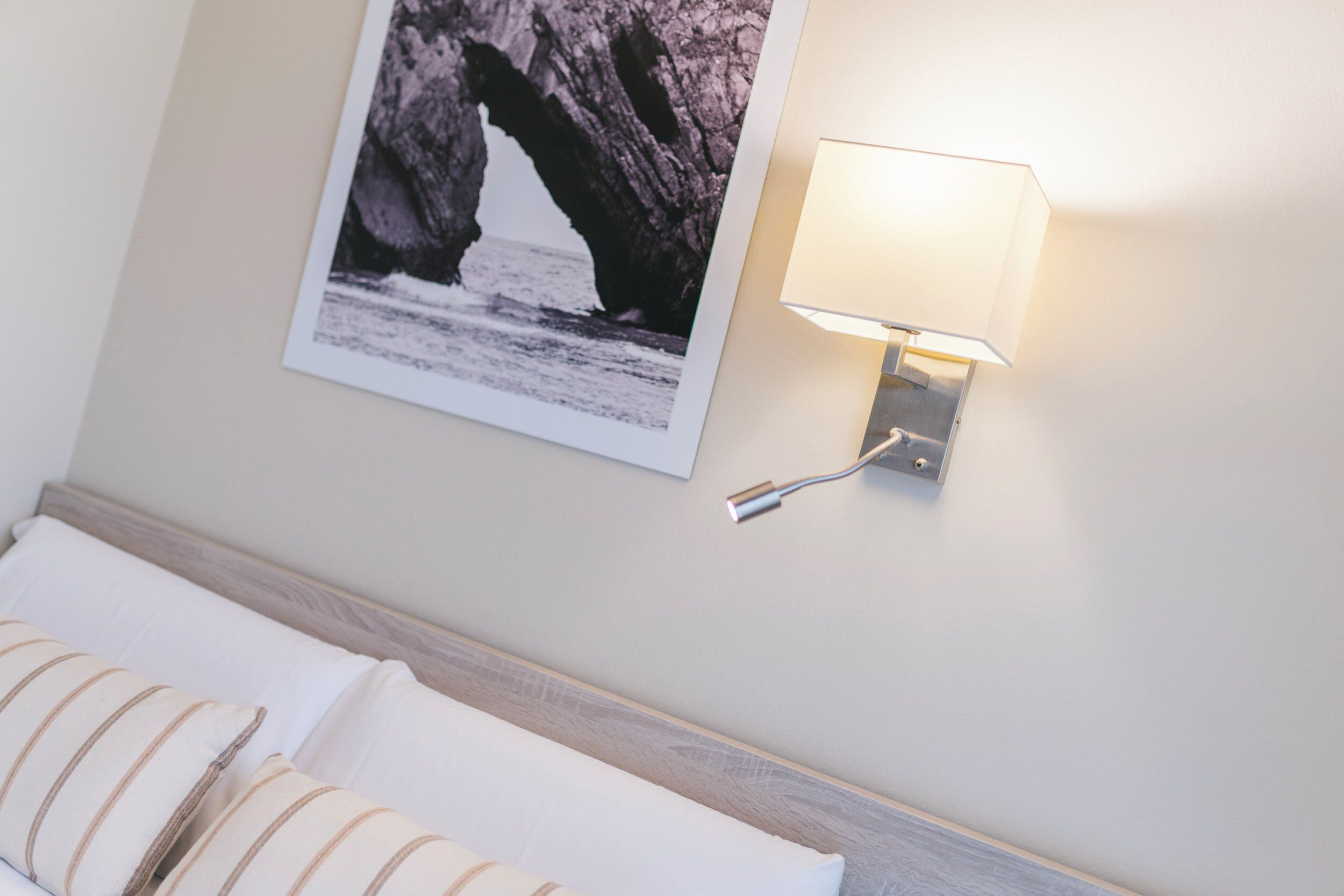 A hotel room with a bed, a wall-mounted lamp, and a framed picture of a coastal rock formation.