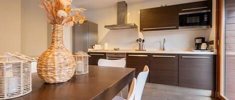A modern kitchen with dark cabinets, a stainless steel range hood, and a microwave. There is a dining table with white chairs and a woven vase on it.
