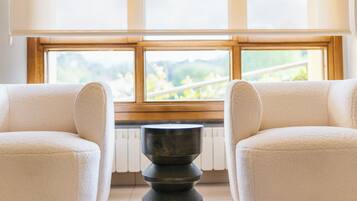 Una habitación con dos sillones beige orientados hacia una ventana con vistas a zonas verdes. Una mesa redonda oscura está en el centro.