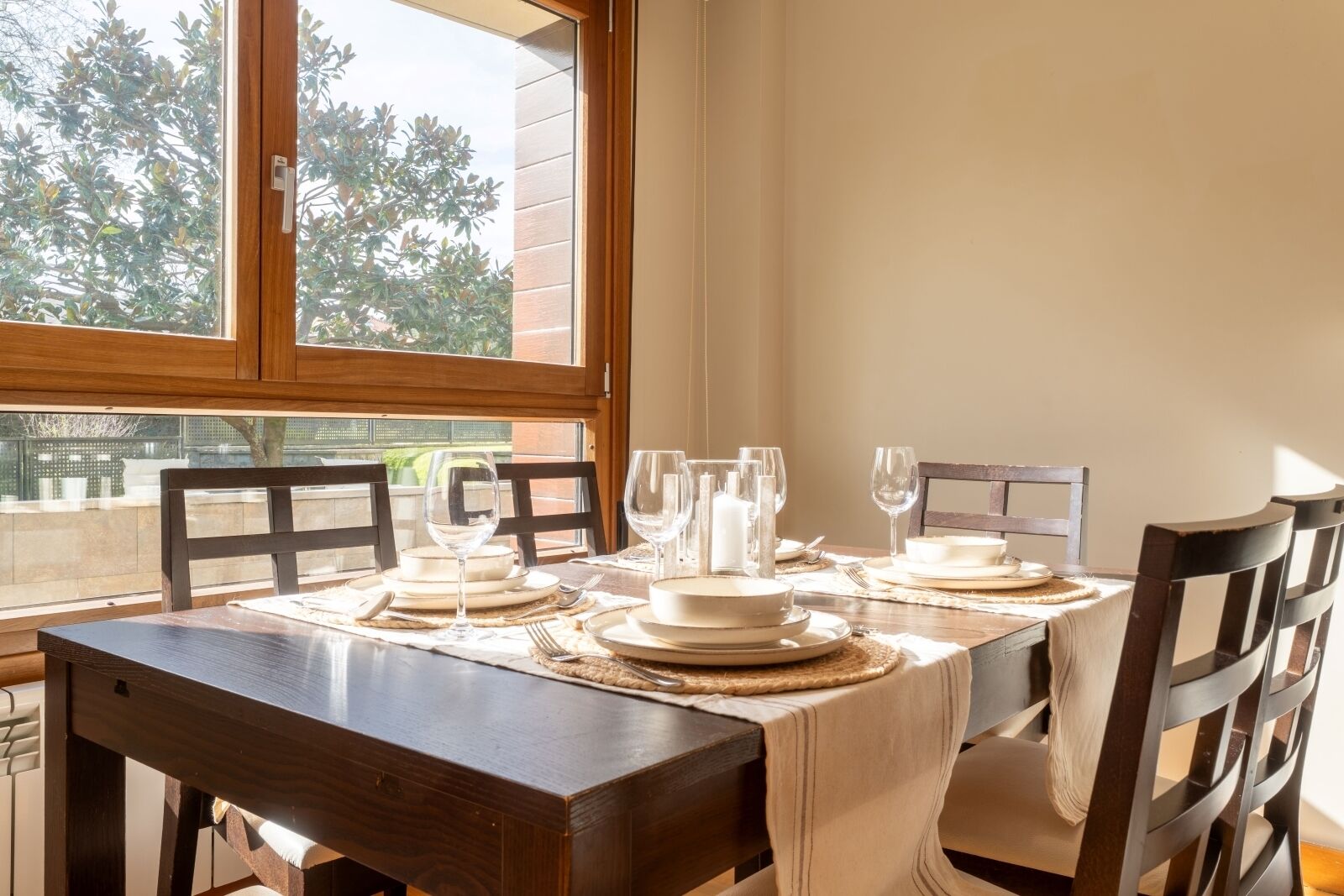 A dining table set for four with white plates, glasses, and a candle, positioned by a window with a view of trees and a fence.
