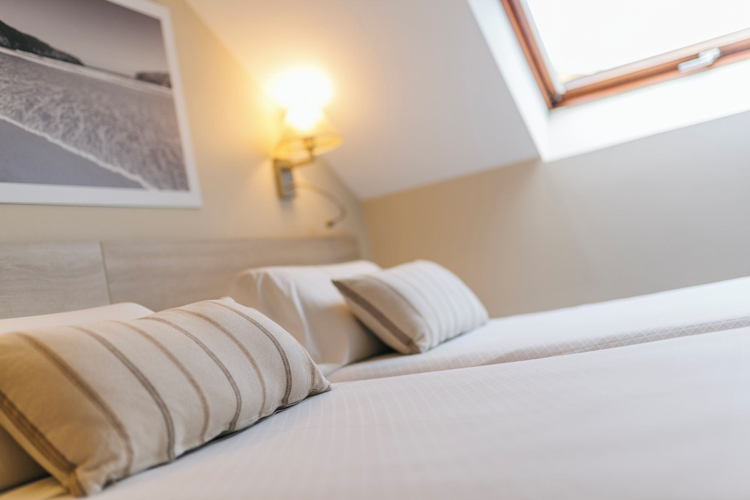 Habitació d'hotel amb dos llits, una taula de nit, una làmpada penjada a la paret i un quadre amb marc sobre el llit.