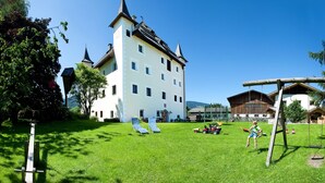 A large white building with a steep roof and a tower, surrounded by a well-maintained lawn with two lounge chairs and a swing set. - Schloss Saalhof (Maishofen)