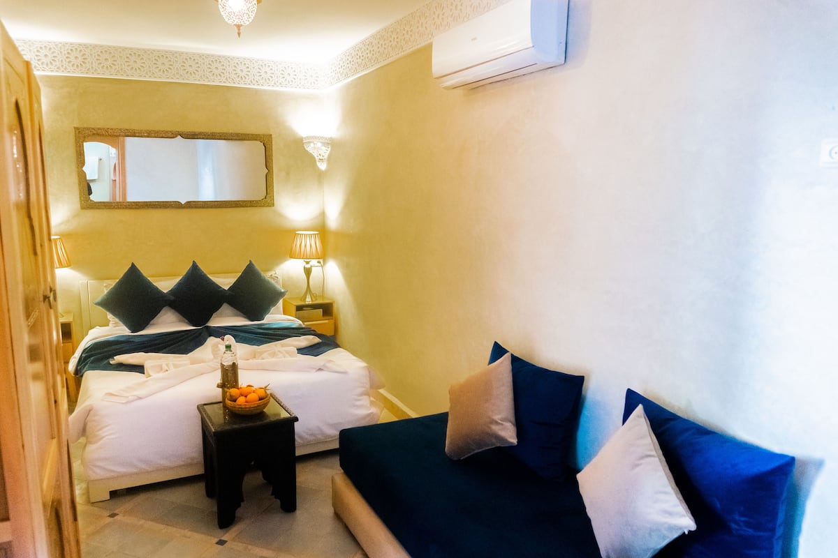 A hotel room with a bed, a bedside table with a fruit bowl, a lamp, and a sofa with pillows.