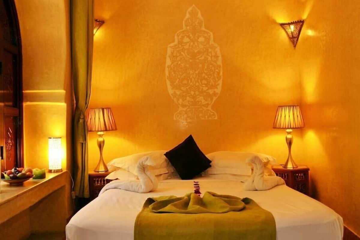 A hotel room with a bed, two bedside lamps, a wall-mounted decorative piece, and a nightstand with a lamp and fruit bowl.