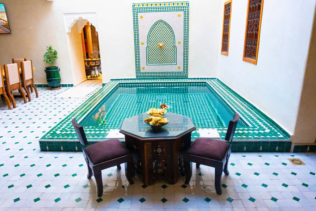 A traditional Moroccan interior with a tiled swimming pool, a wooden table with chairs, and a potted plant.