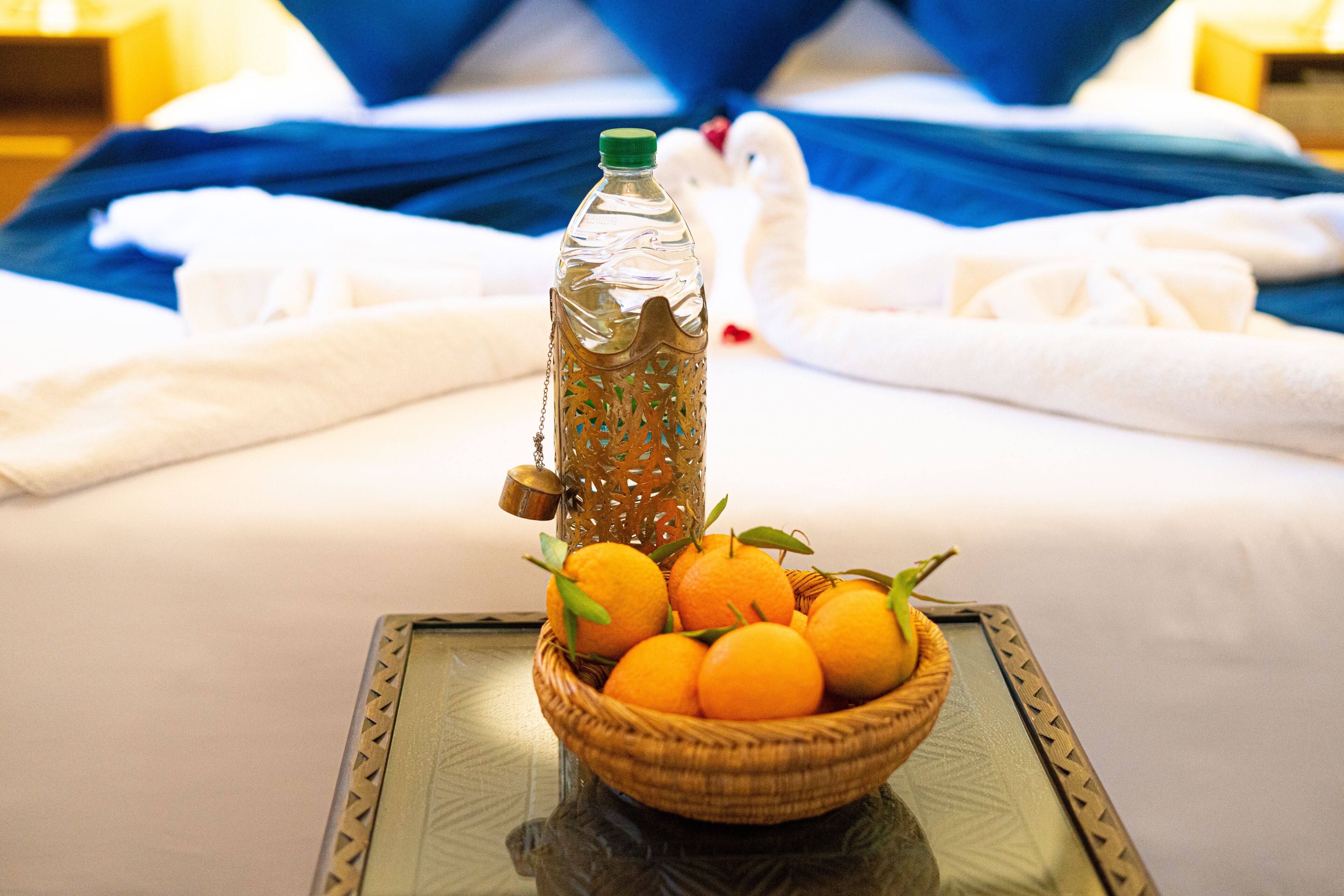 Ein Bett mit blauer Bettwäsche, ein Korbwäscher mit Orangen und eine Glasflasche mit grünem Verschluss auf einem Glastisch.