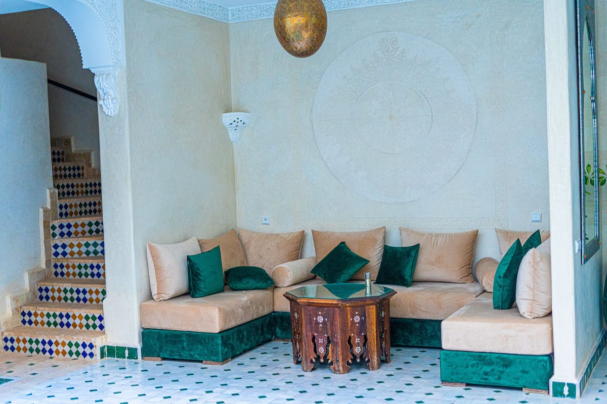 A living room with a beige sofa, green cushions, a glass coffee table, and patterned floor tiles.
