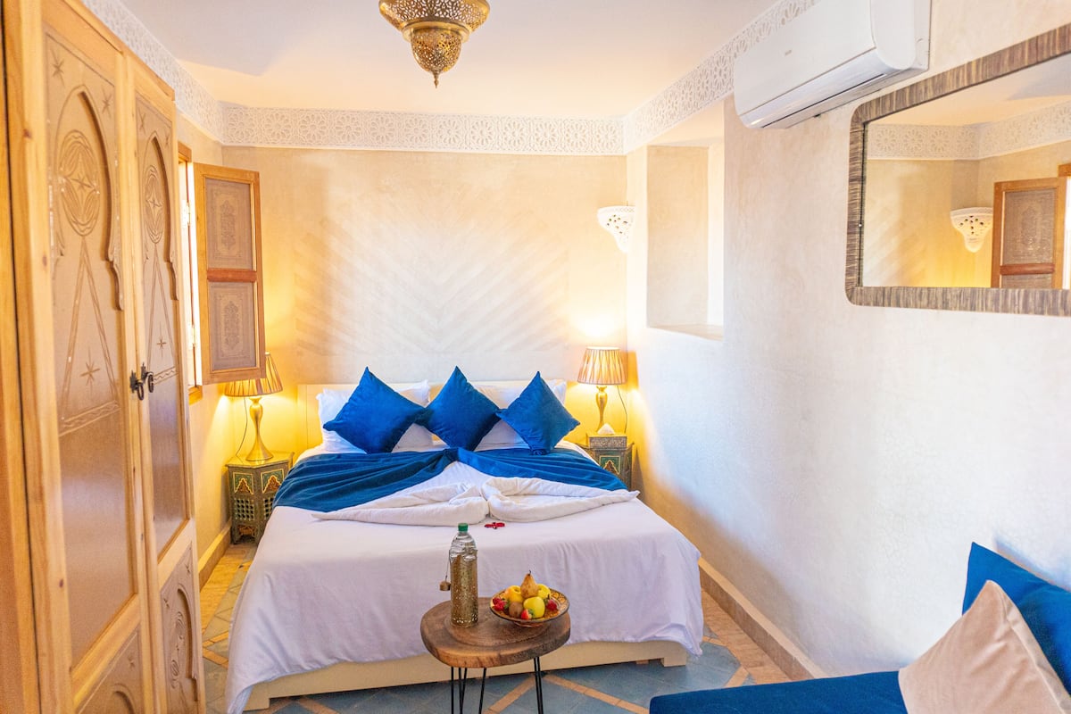 A hotel room with a bed, two bedside lamps, a small table with a bottle and fruit, and a mirror on the wall.