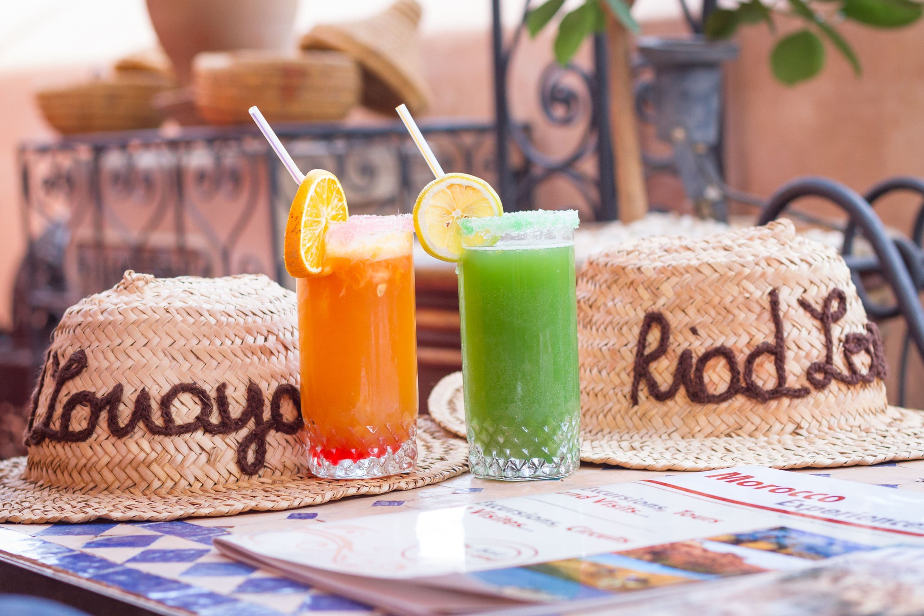 Dos sombreros tejidos, con las palabras "Loujo" y "Roda" inscritas, sobre una mesa junto a dos bebidas en vasos, una anaranjada y otra verde, con pajitas y adornos.
