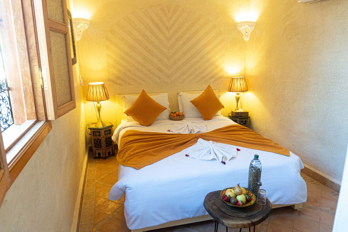 A neatly made bed with a white and orange bedspread, two pillows, and a fruit bowl on a side table.