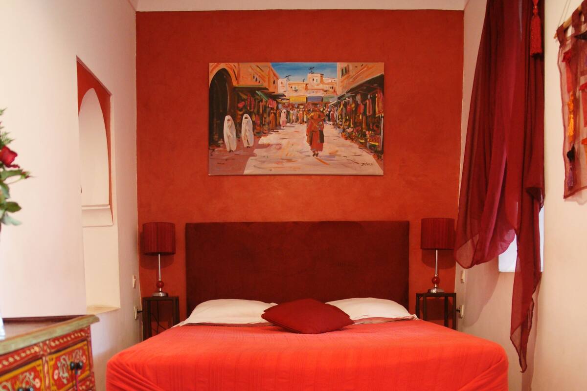 A bedroom with a large bed, a red headboard, two bedside lamps, a red carpet, and a painting on the wall.