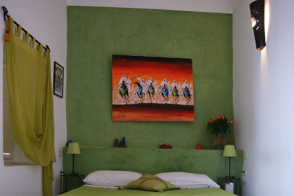 A bed with a green bedspread, two pillows, and a headboard. There is a painting on the wall above the bed, a small table with a lamp, and a window with yellow curtains.
