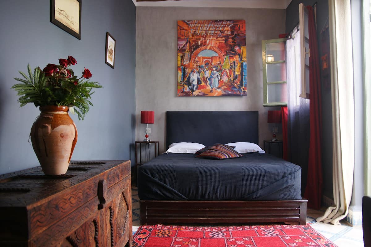 A bedroom with a large bed, a wooden dresser, a vase with flowers, and a colorful painting on the wall.