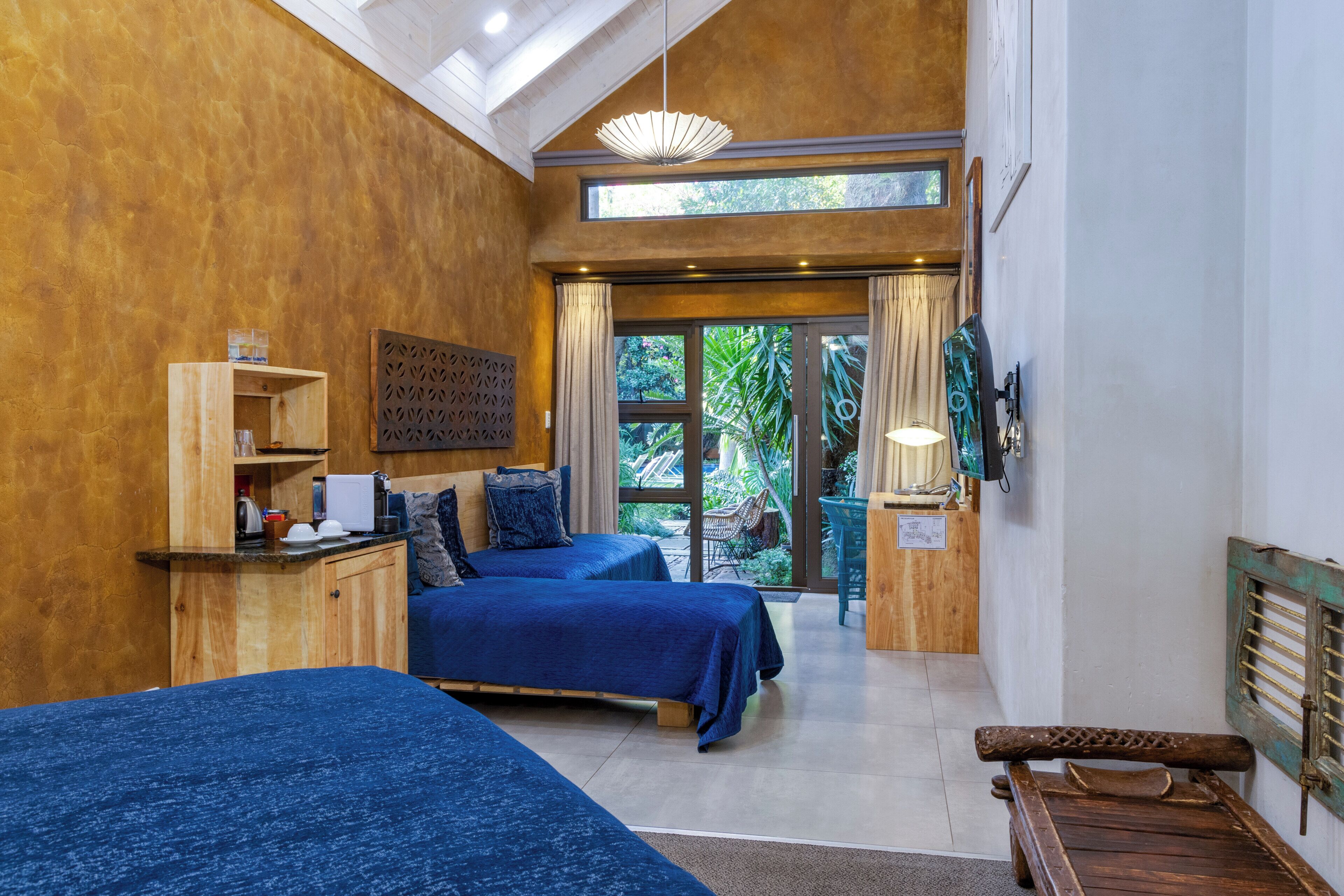 A bedroom with two beds, a wooden bench, a small table with a kettle, and a view of greenery through a large window.
