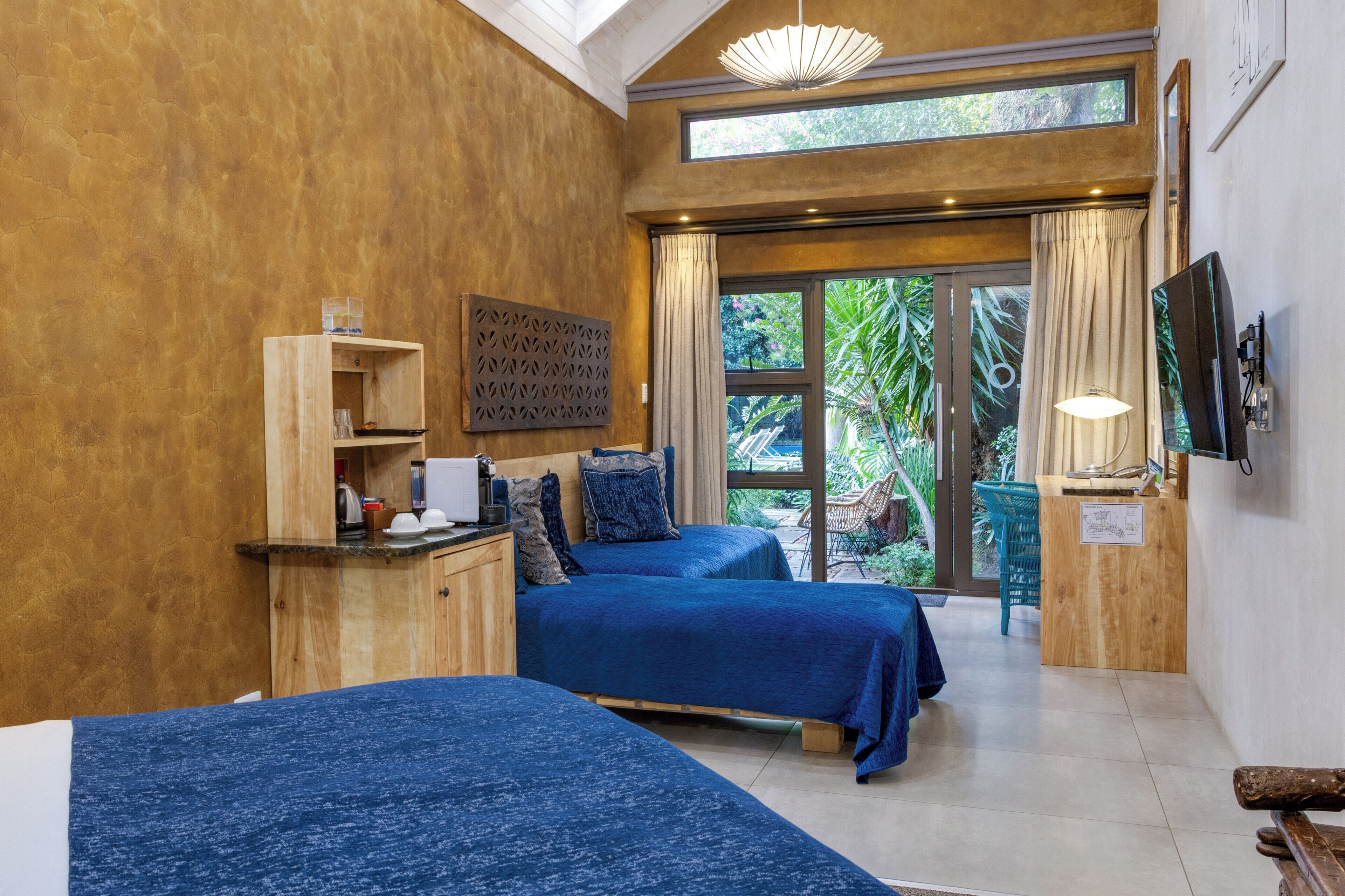 A room with two beds, a wooden desk, a TV, and a large window with a view of greenery.