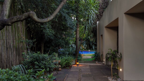 Un sendero de piedra que conduce a una zona de piscina, rodeada de abundante vegetación y una valla de bambú.