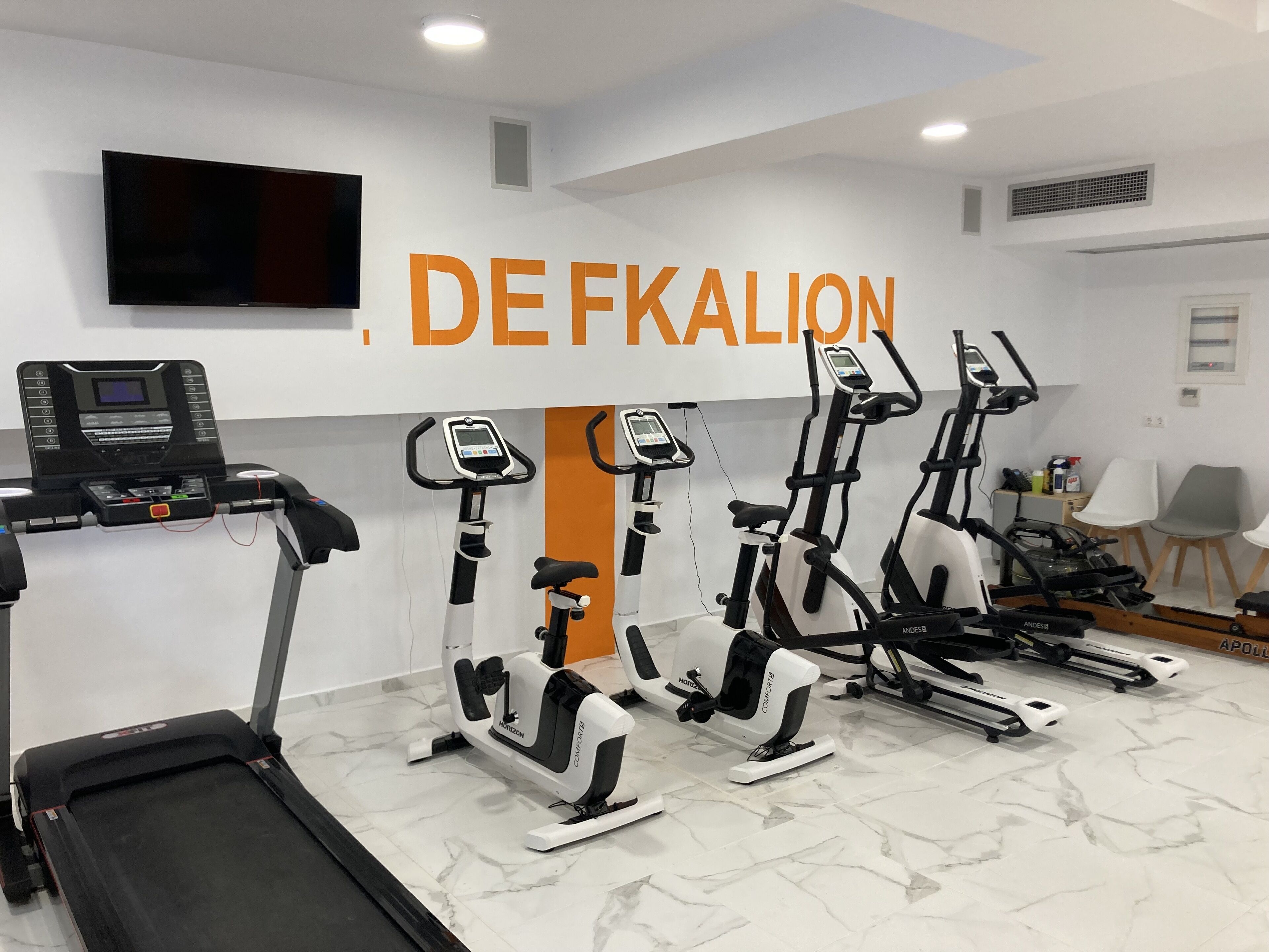 Uma academia com diversos equipamentos de cardio, esteira e televisão presa à parede.