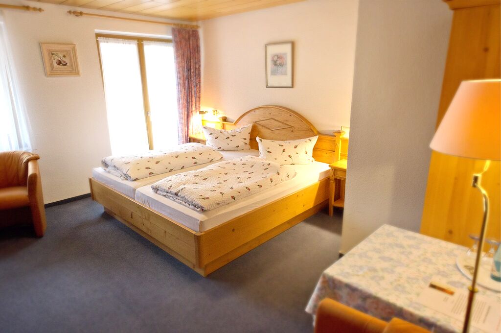 A double bed with a wooden frame, two bedside tables with lamps, a chair, and a small table with a lamp.