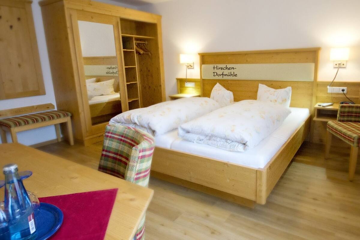 Una cama con estructura de madera y cabecera con la leyenda "Hirschen-Dorfmühle", un banco, una silla y una pequeña mesa con una botella.