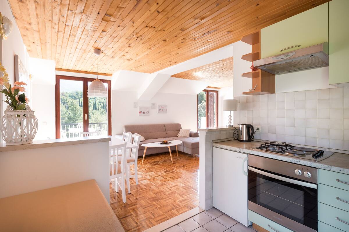 Eine moderne Küche mit Holzbalkendecke, ein Essbereich mit einer Couch und Stühlen sowie ein Blick ins Freie.