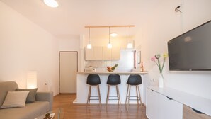 A modern kitchen with white cabinets, a central island with stools, and a flat-screen TV mounted on the wall. - Apartments Karacic (Konavle)